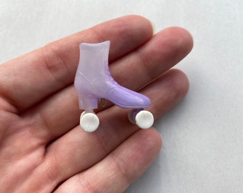 Könnte beinhalten: Ein Miniatur-Rollschuh-Anhänger in Lila- und Weißtönen. Der Skate hat ein klassisches Stiefeldesign mit Schnürsenkeln, einem kleinen Absatz und weißen Rollen. Dieser Anhänger ist ein süßes Accessoire.