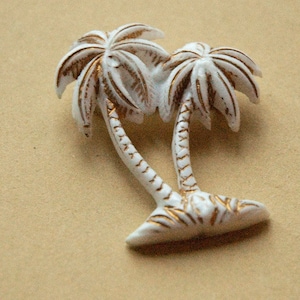 May include: A white and gold palm tree brooch. The brooch depicts two palm trees with white fronds and trunks, highlighted with gold detailing. A tropical-themed accessory.
