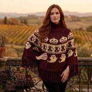 May include: A dark burgundy knit poncho featuring a high turtleneck collar and long fringe trim along the hem. The garment is decorated with a repeating cream-coloured pattern of jack-o'-lanterns, black cats, and ghosts across the chest and shoulders.