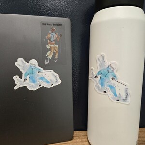 Op de afbeelding: Een witte waterfles en een donkergrijze laptop, beide voorzien van een sticker van een skiër in een blauw jack die door de sneeuw snijdt. De laptop heeft ook een rechthoekige sticker van een wandelaar met de tekst Hike More, Worry Less.
