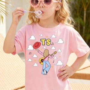 Op de afbeelding: Een lichtroze T-shirt met korte mouwen en een grafische print van een persoon met blond haar die gitaar speelt, een blauwe spijkerbroek draagt en een rode hoed vasthoudt. Het ontwerp bevat witte wolken, muzieknoten en de gele tekst TS.