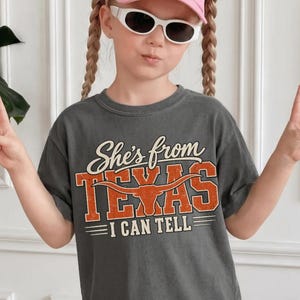 May include: A charcoal gray short-sleeve t-shirt featuring the text She's from TEXAS I CAN TELL in white and orange lettering. The word TEXAS includes a graphic of a longhorn steer skull. The shirt has a crew neckline and a relaxed fit.