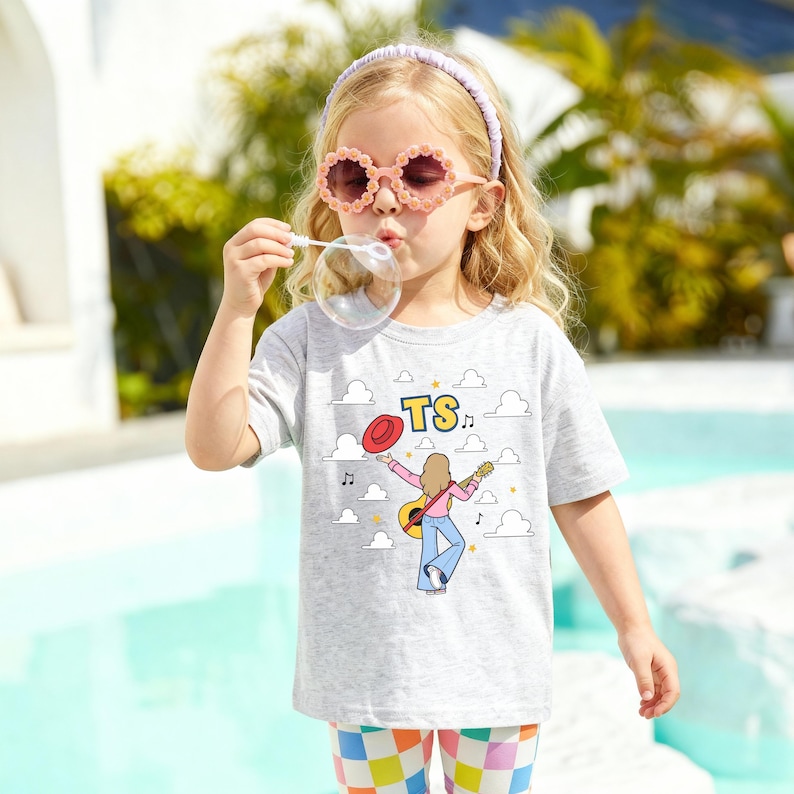 Op de afbeelding: Een lichtgrijs t-shirt met korte mouwen en een grafische print van een persoon die gitaar speelt, een rode hoed, muzieknoten en wolken met de letters TS. Het shirt heeft een ronde hals en een relaxte pasvorm met een gedetailleerde illustratie.