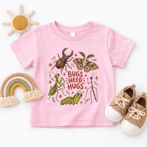 May include: A light pink short-sleeve t-shirt featuring a central graphic of various insects including a beetle, moth, praying mantis, caterpillar, and stick bug, with the text "BUGS NEED HUGS" printed in brown letters surrounded by small star accents.