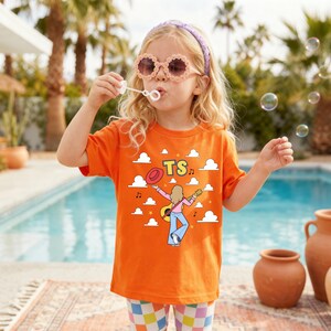 Op de afbeelding: Een oranje T-shirt met korte mouwen en een grafische print van een persoon met lang haar die gitaar speelt, een broek met wijde pijpen draagt en een rode hoed vasthoudt. Het ontwerp bevat witte wolken, muzieknoten en de gele tekst TS.