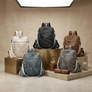 May include: Five backpacks in cream, black, brown, blue-gray, and dark gray. Each bag features a top handle, a front zippered pocket, side pockets, and a wide, patterned shoulder strap with metal grommets. The bags are displayed on various platforms.