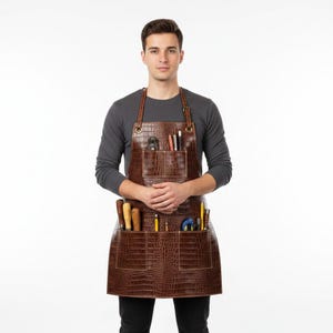 May include: A brown apron with a textured crocodile skin pattern, featuring a single chest pocket and two large lower pockets. The apron includes adjustable straps with metal buckles and holds various tools, including pencils and small hand tools.
