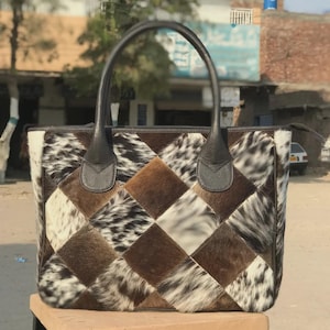 Puede incluir: Un bolso tote con un diseño de patchwork de cuadrados de piel de vaca marrón y blanca. El bolso tiene dos asas de cuero marrón oscuro unidas a la parte superior. El patrón consiste en parches alternos de piel marrón y pelaje moteado.