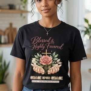 Puede incluir: Una camiseta negra de manga corta con un estampado gráfico de dos manos sosteniendo una rosa rosa y una cruz. El texto dice Blessed & Highly Favored, Grace Upon Grace y bill 4 1:16 -- Of His Fallows We Have All Reviesal.