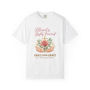 Könnte beinhalten: Ein weißes kurzärmeliges T-Shirt mit einer Grafik von zwei Händen, die eine rosa Rose und ein goldenes Kreuz halten. Über der Grafik steht der Text Blessed & Highly Favored, darunter der Text GRACE UPON GRACE und John 1:16 – Of His Fulness We Have All Received.
