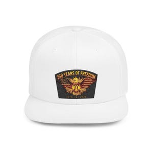 Peut inclure: Une casquette de baseball blanche avec un écusson central représentant un pygargue à tête blanche devant un drapeau américain. Le texte 250 YEARS OF FREEDOM est arqué au-dessus de l'aigle, et les années 1776 et 2026 sont imprimées en dessous.