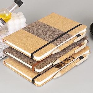 Puede incluir: Una pila de tres cuadernos rectangulares con cubiertas de textura de corcho en varios tonos de beige y marrón oscuro. Cada cuaderno cuenta con una banda elástica horizontal y un bolígrafo de corcho a juego sujeto al lateral.