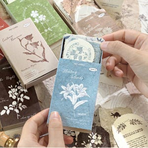 May include: A blue rectangular box labeled Healing Islands containing 30 pieces of decorative paper with floral illustrations. The box features a white lily graphic and text. Surrounding it are other boxes in green, brown, and dark brown with botanical prints and text like Continue Romance and Biologists.