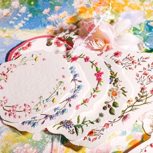 May include: A collection of scalloped-edge paper tags featuring various botanical illustrations. Each tag displays a unique arrangement of colourful flowers, leaves, and berries in shades of pink, blue, green, and yellow on a textured white surface.