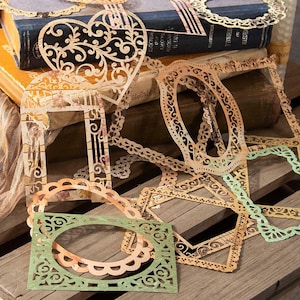 May include: A collection of intricate, laser-cut paper frames and shapes in various geometric and heart designs. These delicate, die-cut paper embellishments feature ornate, lacy patterns in shades of beige, tan, and soft green, layered together.