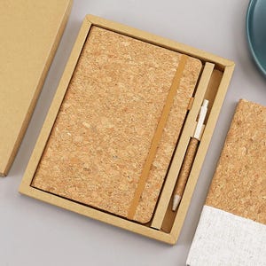 Puede incluir: Un cuaderno rectangular con cubierta de corcho, cierre de banda elástica vertical y un bolígrafo a juego con textura de corcho, presentado dentro de una caja de regalo de cartón color canela. Se ve una tapa de cartón separada y otro cuaderno.