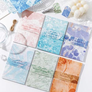 May include: Six rectangular paper packs with varied patterns including pink marble, blue water, purple swirls, light blue textures, green marble, and orange stains. Each pack features a paper band with text such as Shadow Romantic, Shining Sea, Fantasy Wonderland, Like A Dream, Mottled Flecting Time, and Slowness Time.