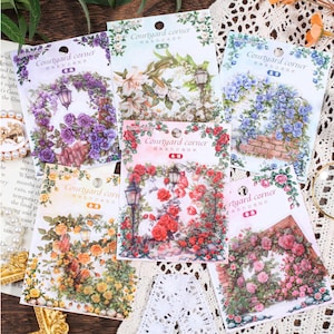 Könnte beinhalten: Sechs rechteckige Aufkleberbögen mit der Aufschrift Courtyard corner, die jeweils Blumenbögen mit Laternen in violetten, weißen, blauen, gelben, roten und rosafarbenen Rosen auf Ziegel- oder Steinstrukturen zeigen, mit Aufhängelöchern oben.