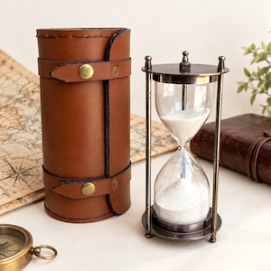 May include: A brass hourglass with white sand positioned next to a cylindrical brown leather case featuring two brass snap closures. The hourglass has a metal frame with a circular top and base, and the leather case is secured with stitched edges.