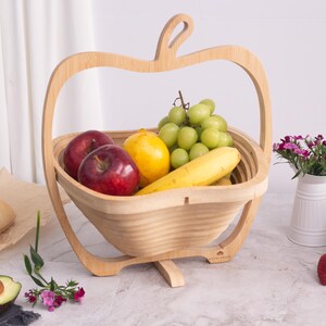 May include: A light-colored wooden fruit basket shaped like an apple with a handle. The basket is filled with two red apples, a yellow lemon, a yellow banana, and a bunch of green grapes. The structure is made of thin, flexible wood slats.