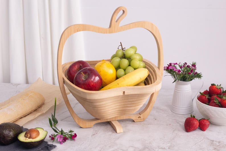 May include: A light-colored wooden fruit basket shaped like an apple with a handle. The basket is filled with two red apples, a yellow lemon, a yellow banana, and a bunch of green grapes. The structure is made of thin, flexible wood slats.