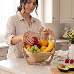 May include: A light-colored wooden fruit basket shaped like an apple, featuring a collapsible design with a central handle. The basket is filled with a variety of fresh produce, including a red apple, a yellow banana, green grapes, and oranges.