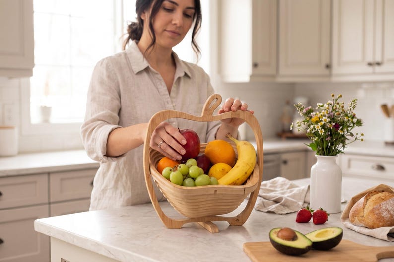May include: A light-colored wooden fruit basket shaped like an apple, featuring a collapsible design with a central handle. The basket is filled with a variety of fresh produce, including a red apple, a yellow banana, green grapes, and oranges.