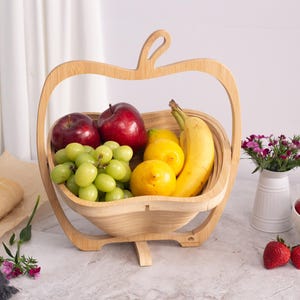 May include: A light-colored wooden fruit basket shaped like an apple, containing red apples, green grapes, yellow lemons, and a banana. Nearby are a baguette, a halved avocado, a white pitcher with small pink flowers, and a bowl of strawberries.