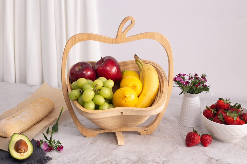 May include: A light-colored wooden fruit basket shaped like an apple, containing red apples, green grapes, yellow lemons, and a banana. Nearby are a baguette, a halved avocado, a white pitcher with small pink flowers, and a bowl of strawberries.