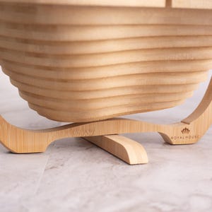 May include: A light brown bamboo fruit basket with a collapsible, tiered, and slatted design. The base features a curved stand with the text Royal House printed on the right side. The structure is made of light-colored wood with visible grain.