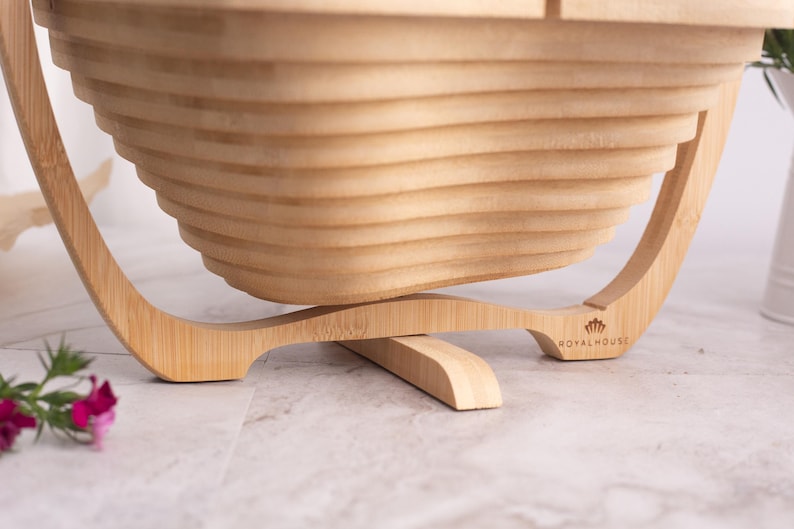 May include: A light brown bamboo fruit basket with a collapsible, tiered, and slatted design. The base features a curved stand with the text Royal House printed on the right side. The structure is made of light-colored wood with visible grain.