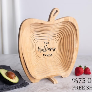 May include: A light-colored wooden collapsible basket shaped like an apple with a stem at the top. The center of the basket is engraved with the text The Williams Family. The basket is displayed in an open, expanded position on a flat surface.