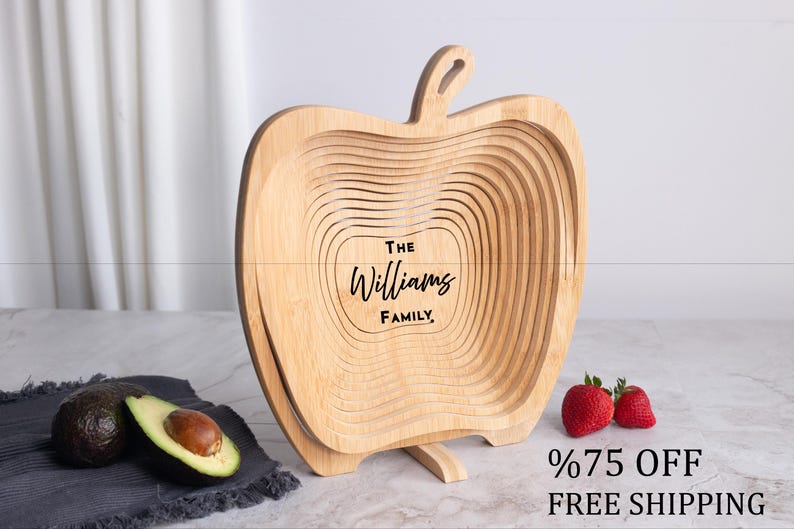 May include: A light-colored wooden collapsible basket shaped like an apple with a stem at the top. The center of the basket is engraved with the text The Williams Family. The basket is displayed in an open, expanded position on a flat surface.
