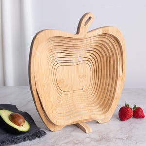 May include: A light brown bamboo fruit basket shaped like an apple with a collapsible, spiral-cut design. The basket features a central stem-like handle at the top and a flat base for stability, showcasing the natural wood grain and texture.