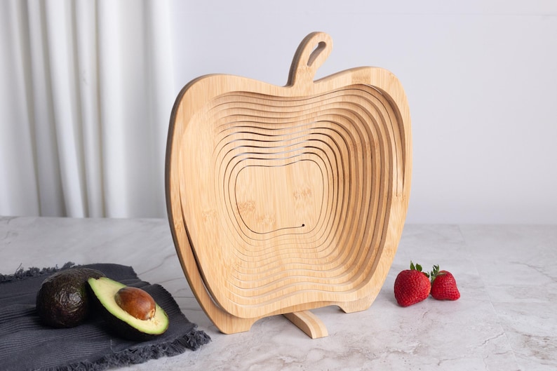 May include: A light brown bamboo fruit basket shaped like an apple with a collapsible, spiral-cut design. The basket features a central stem-like handle at the top and a flat base for stability, showcasing the natural wood grain and texture.
