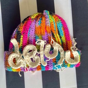 Peut inclure: Un bonnet en crochet multicolore présentant un mélange vibrant de fils rose néon, orange, bleu et violet. Le bonnet est orné de lettres en crochet de couleur crème formant le mot "delulu" sur le devant, avec des fils qui dépassent.