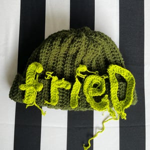 Peut inclure: Un bonnet crocheté vert olive avec le mot "fried" appliqué en fil vert citron vif sur le devant. Les lettres sont cousues sur la texture en tricot côtelé du bonnet, avec des fils lâches visibles à la base des lettres.