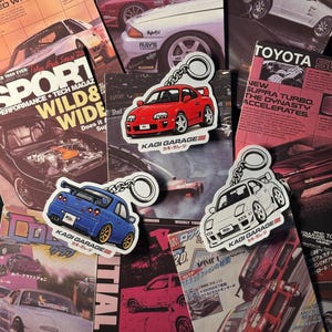 May include: Three car-shaped keychains featuring a red Toyota Supra, a blue Nissan Skyline GT-R, and a white Mazda RX-7, each with a silver metal ring. The keychains are displayed over a collage of vintage automotive magazine covers.