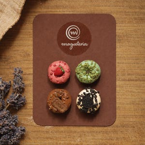 May include: A brown rectangular display card featuring the Magneteria logo and four miniature donut magnets. The donuts include a pink one with a strawberry, a green one with sprinkles, a brown one with a cookie piece, and a white one with dark crumbles.