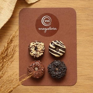 Może przedstawiać: Cztery miniaturowe magnesy w kształcie pączków przymocowane do brązowej prostokątnej karty z okrągłym logo i napisem "magneteria". Pączki są ozdobione posiekanymi orzechami, białymi paskami lukru, kawałkami czekolady oraz ciemną posypką.