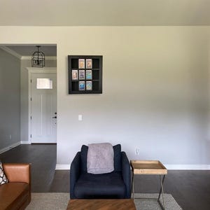 May include: A black square wall-mounted display case with nine individual compartments, each containing a rectangular trading card in a protective plastic slab. The display case is mounted on a plain light-coloured wall above a dark blue armchair.