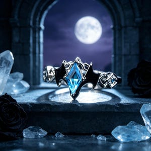 Puede incluir: Un anillo de tono plateado con una piedra preciosa azul central en talla marquesa, flanqueada por dos motivos de alas de murciélago negros. La banda muestra un intrincado patrón geométrico con pequeños detalles en azul.