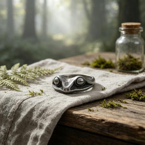 May include: A silver-toned metal ring shaped like a frog head with prominent, detailed eyes and a smooth, curved band. The ring features a weathered, dark finish that highlights the textured contours of the frog's face and its closed eyelids.