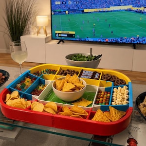 May include: A multi-compartment snack serving tray designed like a soccer field, featuring a red, yellow, and blue frame. It holds various snacks including tortilla chips, popcorn, pretzels, candies, and Snickers bars in separate sections.