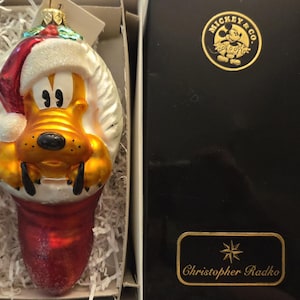 May include: A glass Christmas ornament featuring Pluto wearing a red and white Santa hat, emerging from a red stocking. The ornament is presented in a box with a black lid labeled Mickey & Co. and Christopher Radko in gold lettering.