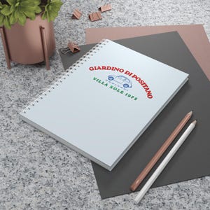 Peut inclure: Un carnet à spirales blanc présentant un graphisme avec le texte rouge GIARDINO DI POSITANO, le contour bleu d'une voiture ancienne et le texte vert VILLA SOLE 1975. La couverture est sobre et met en valeur le motif imprimé de manière claire.