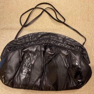 May include: A black shoulder bag with a soft, gathered texture and a long, thin strap. The front features several elongated, leaf-shaped panels with a glossy, textured snakeskin-like pattern, contrasting with the matte finish of the main body.