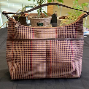 May include: A hobo-style shoulder bag featuring a brown and tan houndstooth pattern with thin red vertical stripes. The bag includes a dark brown leather handle, a top zipper closure, and a horizontal exterior pocket across the front panel.