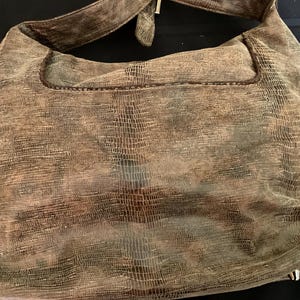 May include: A brown hobo-style shoulder bag featuring a textured, reptilian-inspired embossed pattern across the exterior surface. The bag includes a single wide shoulder strap, a top opening with stitched detailing, and visible metal feet at the base.