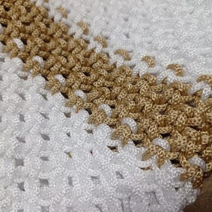 Puede incluir: Un art&iacute;culo textil con un cord&oacute;n blanco y dorado met&aacute;lico tejido en un patr&oacute;n de nudos repetitivos. El dise&ntilde;o consiste en una banda ancha de nudos blancos seguida de una franja diagonal de nudos dorados, creando una superficie de rejilla.